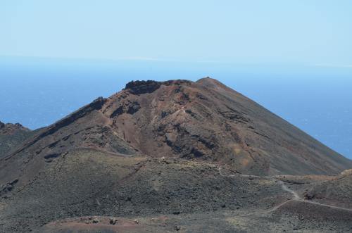 A volcano seen from a distance. There is blue sky and blue sea in the distance. The volcano is not erupting. It is covered in vegetation. The view is into a large hollow caused by a previous eruption.