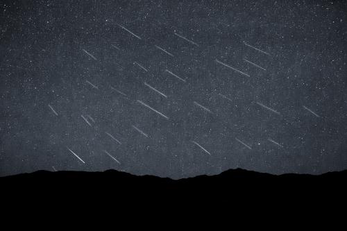 Night sky view of a meteor shower, with multiple bright streaks of light crossing the sky, set against a backdrop of stars and a dark silhouette of mountains in the foreground.