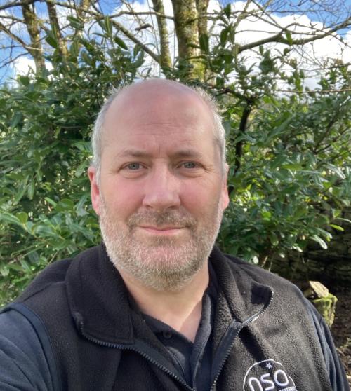 A portrait photo of a man with a beard looking forwards. There are some green brushes and trees in the background.