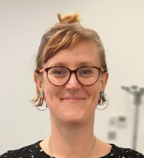 A portrait photo of a woman. She has short blonde-brown hair that is tied into a small bun, and a small side fringe. She is wearing glasses and gold, hooped earrings.