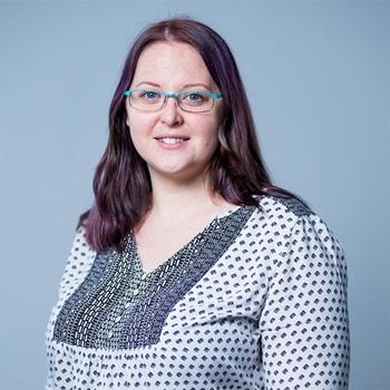 A portrait photo of a woman. She has red-purple, shoulder length hair and is wearing glasses with a light blue frame.