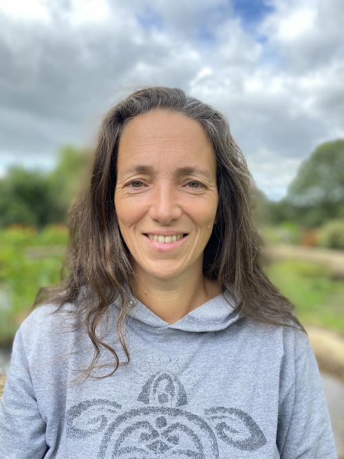 A portrait photo of a woman with shoulder length, brown hair. She is facing forwards and wearing a grey hoodie. There is a green field in the background with a few trees around the edges. There are white clouds in the sky.