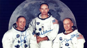 The Apollo 11 crew in their spacesuits but not wearing space helmets. They are shown in front of a large image of the Moon. Neil Armstrong is seated on the left. Michael Collins is standing in the centre. Buzz Aldrin is seated on the right.