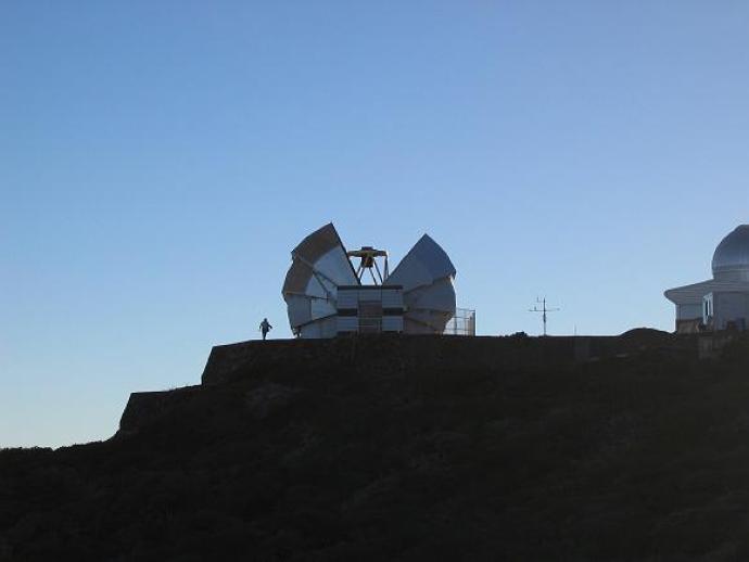 A small, circular building on top of a mountain. The dome is open at the top, as if peeled back slightly, revealing a telescope that is pointed upwards.