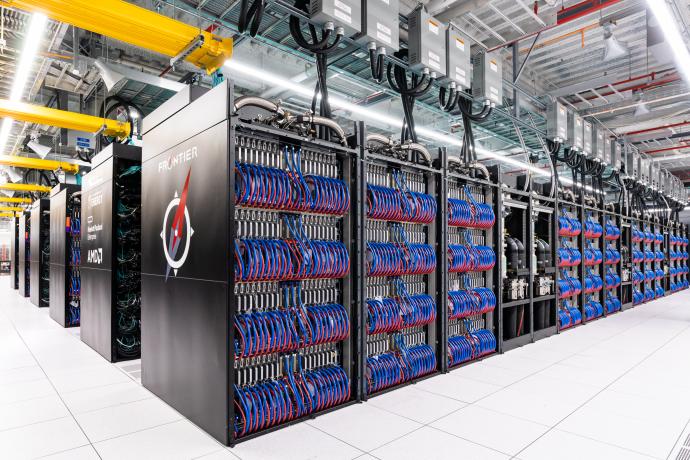 Rows of black cabinets housing the Frontier supercomputer, with blue and red cables neatly organized along the sides. The environment features a clean, industrial look with overhead yellow cable trays and visible ventilation systems