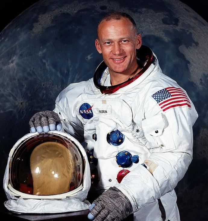 A portrait of astronaut Buzz Aldrin in his NASA spacesuit, smiling and posing with his helmet in one hand. The background features the moon, highlighting his role as part of the Apollo 11 mission