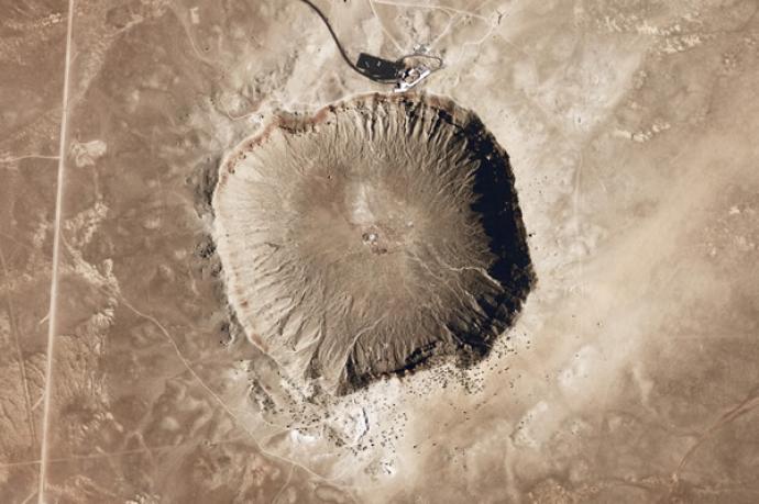 Overhead view of a large meteor crater in a desert landscape, with a small building and road at the edge of the crater