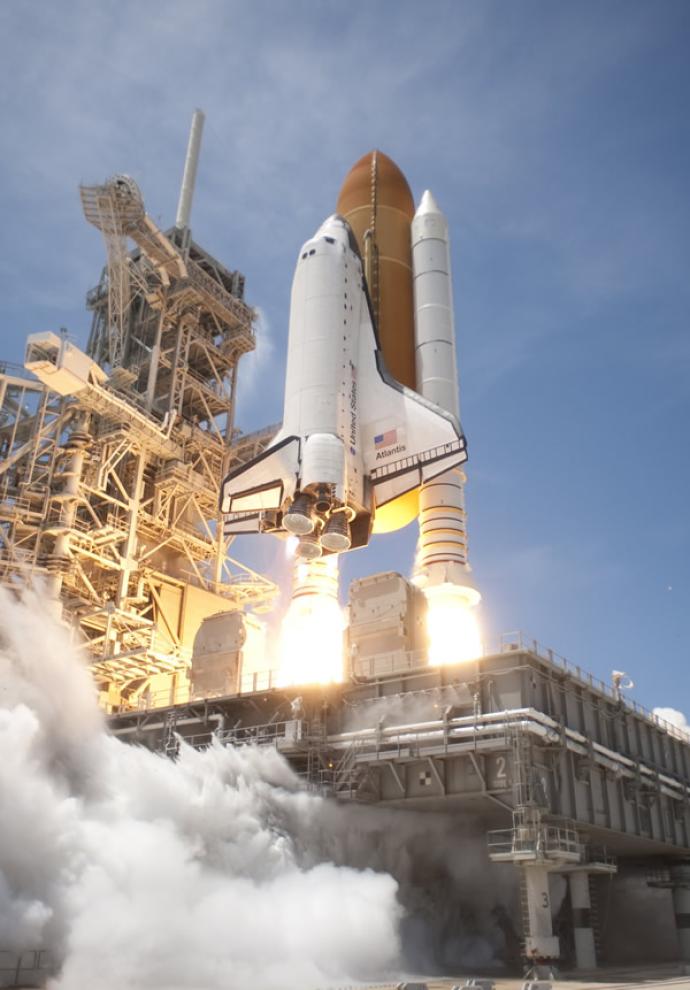 Space Shuttle Atlantis launching from the pad, with smoke and flames billowing from the engines as it ascends into the sky