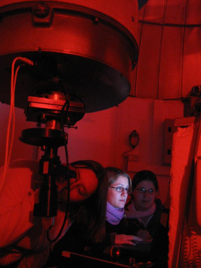 Three people working in a dimly lit observatory, illuminated by red light, as one person looks through a telescope while the others operate equipment