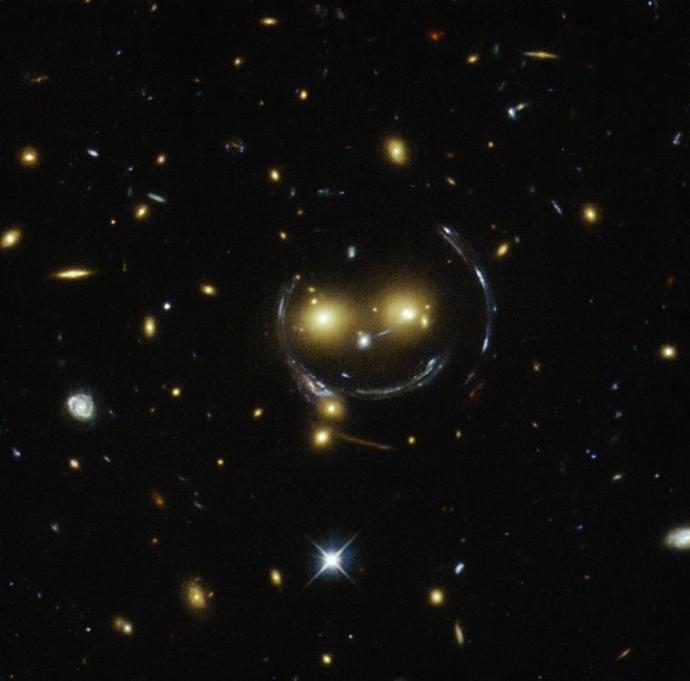 An image showing many galaxies within a cluster. Towards the centre, the light has curved, appearing to form the outline of a face and a mouth smiling whilst two bright galaxies look like the eyes.