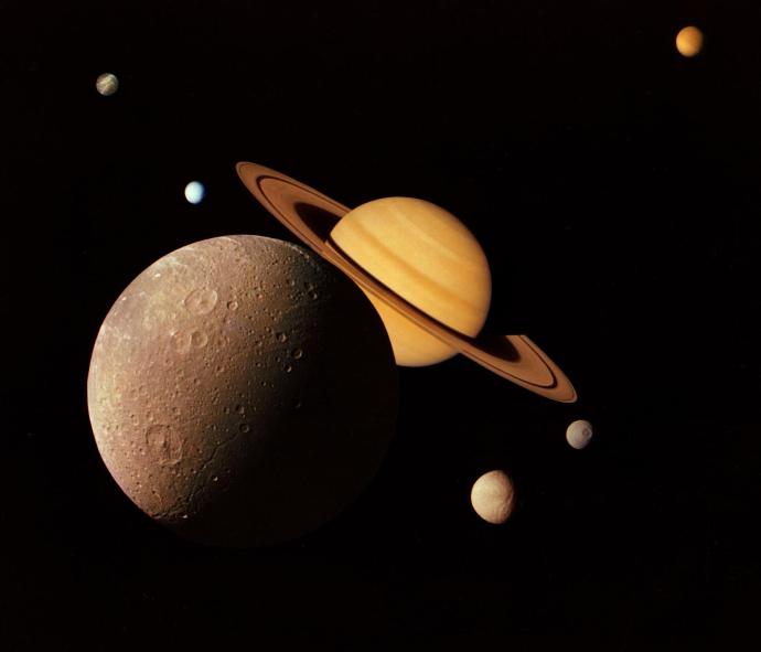 Image of Saturn with its rings, surrounded by several of its moons of varying sizes and colors, against a dark background